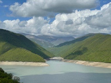 Bulutlu bir gökyüzünün altında dağlarla çevrili sakin bir göl manzarası.