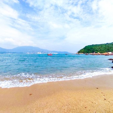 Arka planda dağlar olan kumlu bir sahilde yumuşak mavi deniz suyu dalgalarının çarpıcı görüntüsü.