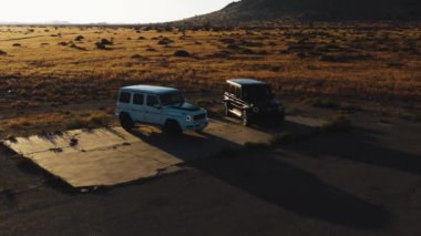 Güneş batarken çöle park etmiş siyah beyaz Mercedes G Wagon 'un insansız hava aracı görüntüsü.