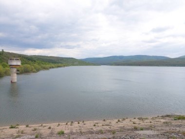 Bulutlu bir gökyüzünün altında ağaçlar ve dağlarla çevrili bir su deposu.