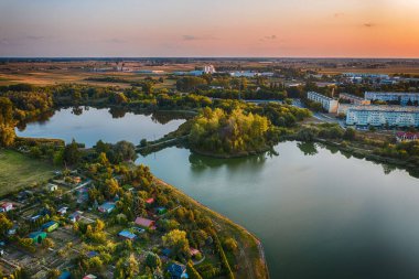 Yeşil bir manzarayla çevrili güzel bir gölün insansız hava aracı görüntüsü. Bulutlu gökyüzünün altında gün batımlı küçük evler.