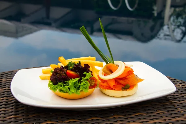A beautiful shot of a salmon burger, lettuce and tomato burger and fries on a white plate