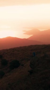 Kırsal tepeler ve su üzerinde parlayan günbatımı gökyüzünün dikey görüntüsü.