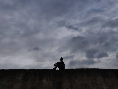 Binanın tepesinde oturan yalnız bir erkeğin silueti bulutlu bir gökyüzüne bakıyor.
