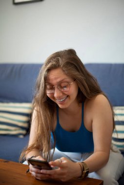 Beyaz tenli sarışın bir kadın evindeki koltukta cep telefonuyla video görüşmesi yapıyor..