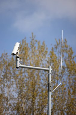 Mavi gökyüzüne karşı gri bir direkteki sokak güvenlik kamerasının dikey odak noktası ve yeşil ağaç arkaplanı.