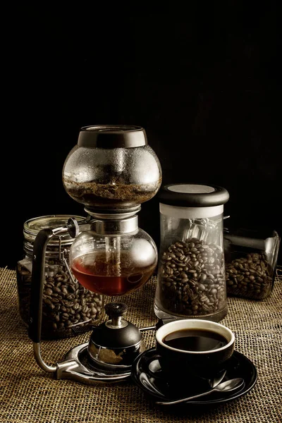 A vertical shot of a cup of coffee, coffee maker, and coffee beans in a jar on a black background