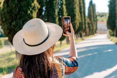 Şapkalı genç bir kadın cep telefonuyla Toskana 'daki güzel bir yolun fotoğraflarını çekiyor.