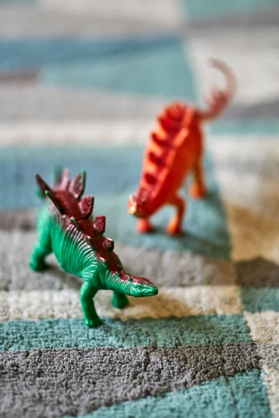 A closeup shot of a green stegosaurus dinosaur toy on the carpet in bright light with a red dinosaur toy in the blurred background