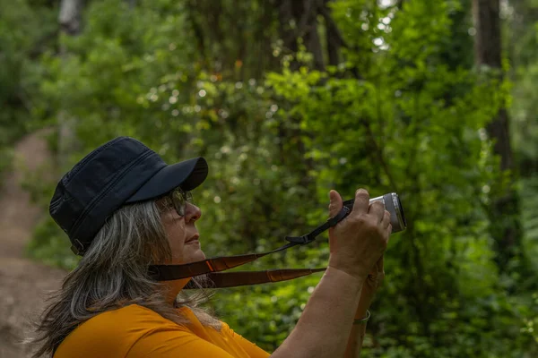 Beyaz saçlı, şapkalı ve turuncu tişörtlü bir kadın ormanda fotoğraf çekiyor.