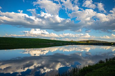 Mavi parlak gökyüzünün altında yeşil tarlalarla çevrili bir nehir manzarası.