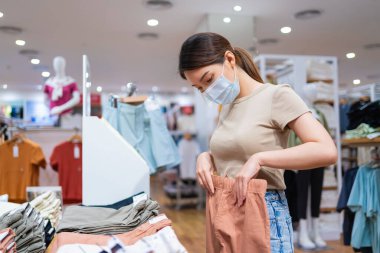 Koruyucu yüz maskesi takan Asyalı bir kadın mağazadan kıyafet alıyor.