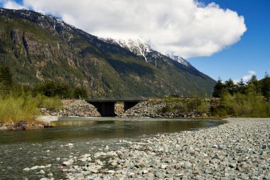 Uzak bir yoldaki bir köprünün manzarası ve arkasındaki karlı dağlar sığ bir nehir yatağının ardında..