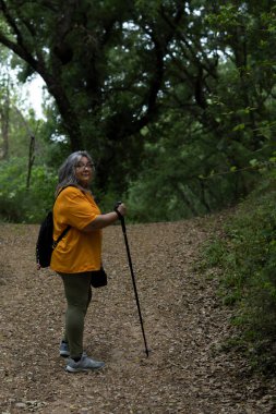 Yürüyüş yolunda yürüyüş yapan beyaz saçlı, bastonlu, sırt çantalı ve turuncu tişörtlü kadın.