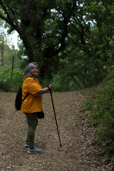 Yürüyüş yolunda yürüyüş yapan beyaz saçlı, bastonlu, sırt çantalı ve turuncu tişörtlü kadın.