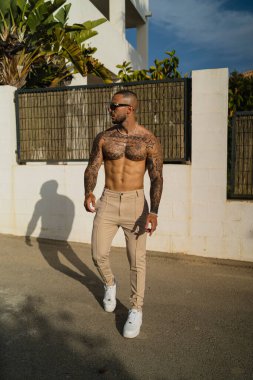 A vertical shot of a young tattooed shirtless man in beige pants and white sneakers
