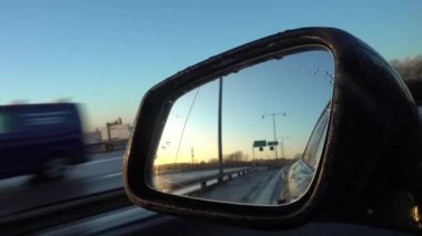 A closeup of a car's side mirror reflecting the highway at sunset