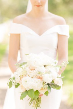 Beyaz bir elbise giymiş, elinde bir buket beyaz gül ve yeşil yaprak tutan bir gelinin dikey görüntüsü.