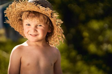A blond Caucasian little boy smiling wearing a straw hat in summer