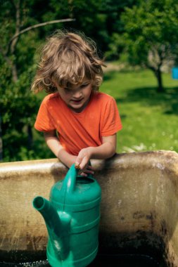 A vertical shot of a cute Caucasian boy collecting water into a green watering can in a garden