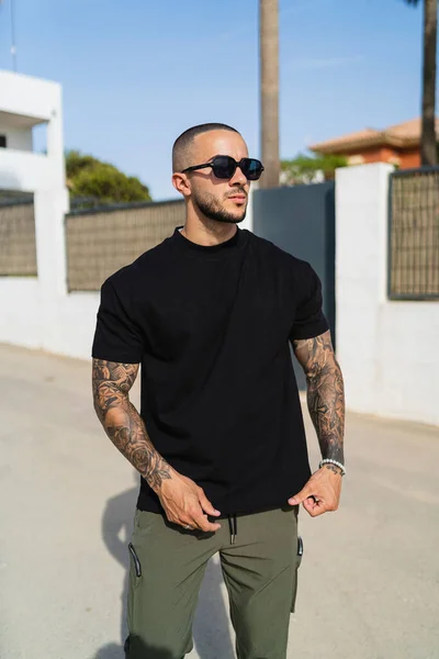 A vertical shot of a young tattooed man wearing a black t-shirt and green pants