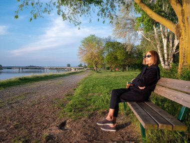 Yetişkin bir kadın. Bacak bacak üstüne atmış, baharda göl kenarında bir bankta oturuyor.