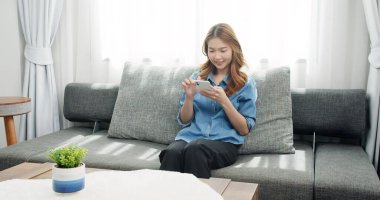 Asyalı kadın kanepede dinleniyor ve akıllı telefonuyla sosyal medyada geziniyor. Mutlu kız erkek arkadaşına mesaj atıyor..