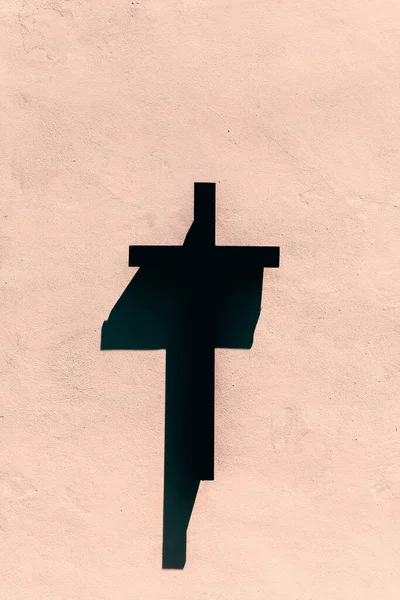 A black wooden cross hanging on a pink exterior wall of a building in bright sunlight