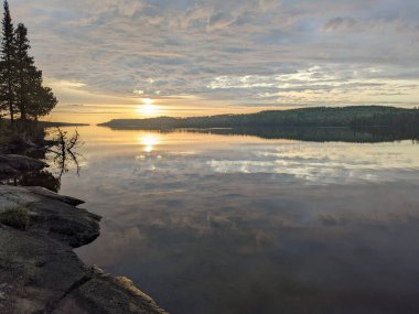 Gün batımında bulutlu bir gökyüzünün güzel yansımasıyla çevrili bir gölün manzarası.