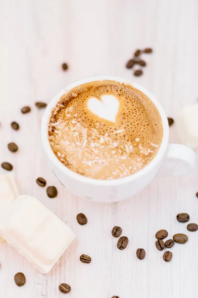 A vertical shot of a cup of coffee with a heart foam surrounded by coffee beans and white chocolate