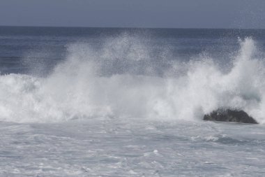 Büyük köpüklü dalgalar yazın güneşli bir gününde deniz kıyısındaki kayalara vuruyor.