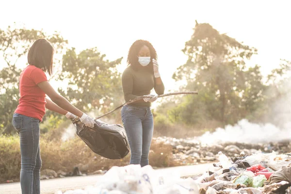 A Nigerian women cleaning up together a dump site with trees and smoke in the background in Nigeria