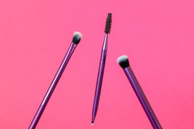 A pair of purple makeup brushes and an eyelash brush on a pink background