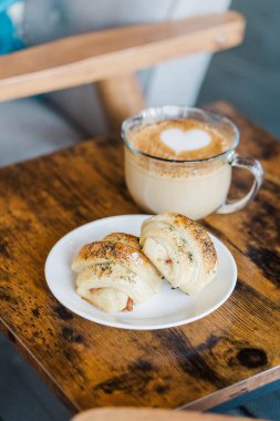 Yüzeyinde kalp tasarımı olan bir kahvenin dikey görüntüsü ve küçük bir masada beyaz bir tabakta hamur işi atıştırmalıkları.