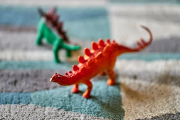 A closeup shot of a red stegosaurus dinosaur toy on the carpet in bright light with a green dinosaur toy in the blurred background