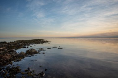 A body of water near the rocky shore reflecting the light cloudy sky