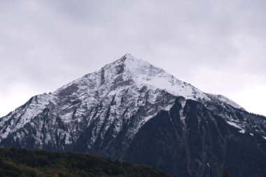 Kasvetli gökyüzünün altında karla kaplı kayalık bir dağın manzarası.