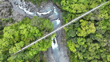 Güneşli bir günde, vahşi bir ormanda bir dağ nehrinin üzerindeki yaya köprüsünün hava görüntüsü.