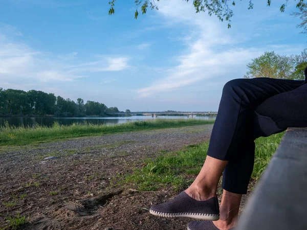 Woman with her legs crossed sitting on a park bench near a lake in springtime