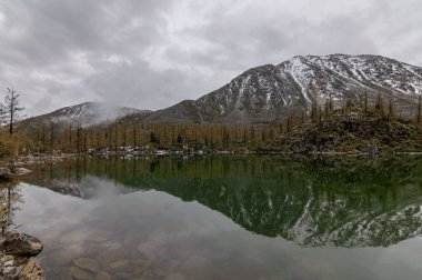 Karlı dağların sakin manzarası bulutlu bir havada göle yansıyor.