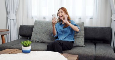 Evde akıllı telefonlu Asyalı genç bir kadının portresi. Çekici kız kanepede oturmuş, rahat bir yerde arkadaşlarıyla sohbet ediyor.