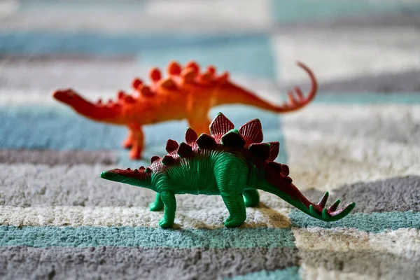 A closeup shot of a green stegosaurus dinosaur toy on the carpet in bright light with a red dinosaur toy in the blurred background