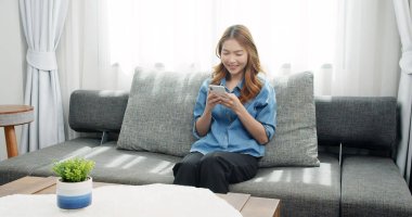 Asyalı kadın kanepede dinleniyor ve akıllı telefonuyla sosyal medyada geziniyor. Mutlu kız erkek arkadaşına mesaj atıyor..