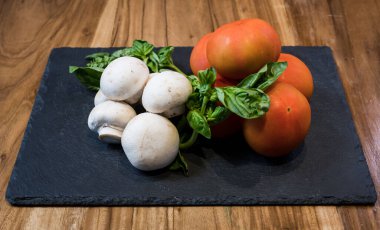 A closeup of fresh mushrooms, tomatoes and basil on a black board on a wooden table
