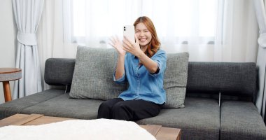 Evde akıllı telefonlu Asyalı genç bir kadının portresi. Çekici kız kanepede oturmuş, rahat bir yerde arkadaşlarıyla sohbet ediyor.