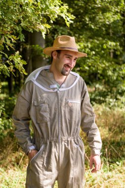 European beekeeper with black hair and beard with protective suit and straw hat has good mood on the edge of forest
