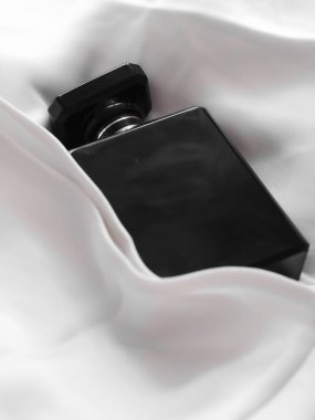 Lüks bir siyah parfüm şişesinin dikey görüntüsü beyaz bir kumaşın üzerinde yatar.