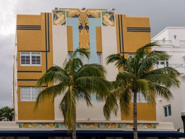 An Art Deco Building ochre or mustard-colored with colorful murals in the middle and palm trees in front in Miami Beach
