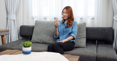 Evde akıllı telefonlu Asyalı genç bir kadının portresi. Çekici kız kanepede oturmuş, rahat bir yerde arkadaşlarıyla sohbet ediyor.