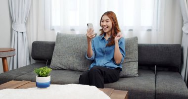 Evde akıllı telefonlu Asyalı genç bir kadının portresi. Çekici kız kanepede oturmuş, rahat bir yerde arkadaşlarıyla sohbet ediyor.